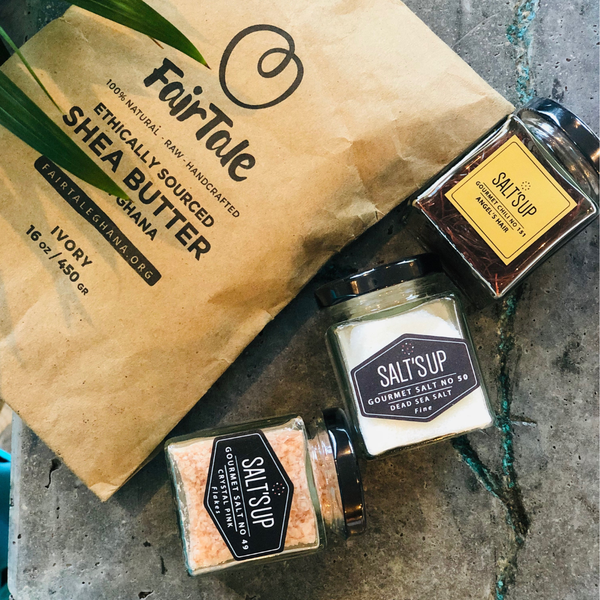FairTale GHANA raw Shea butter pouch beside three jars of gourmet sea salt on a stone surface