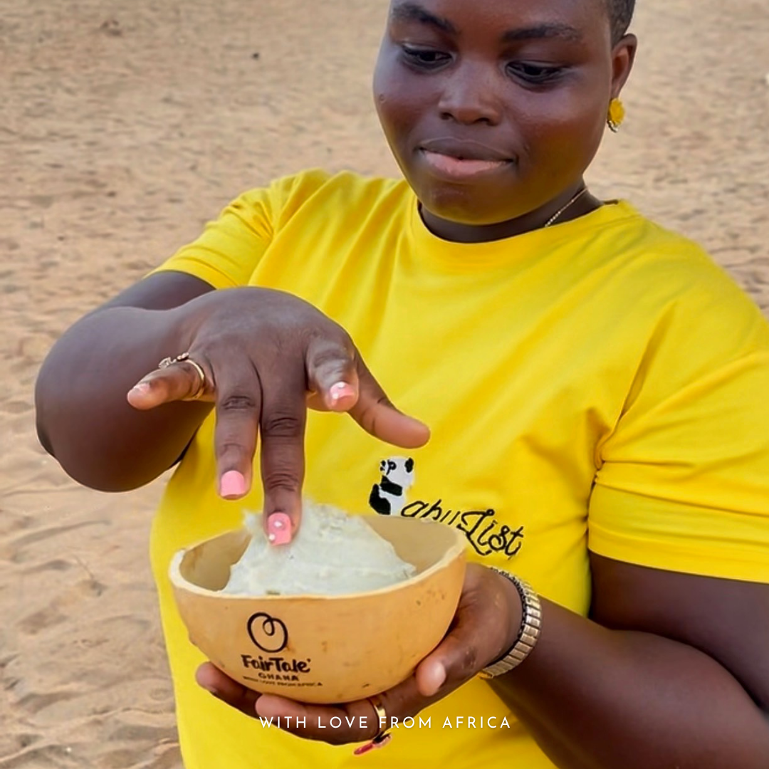 A person wearing a bright yellow t-shirt with a black panda logo and 'chibiLife' text, holding a calabash bowl filled with raw shea butter outdoors on a sandy ground. The calabash bowl also features the brand 'FairTale GHANA'
