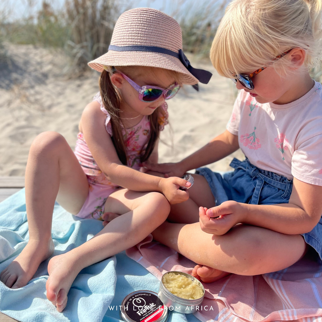 Two young girls sitting on a blue blanket on a sandy beach, applying a FairTale GHANA Shea butter from a small round tin. One girl wears a pink straw hat with a dark ribbon, and the other wears a white t-shirt and denim shorts.